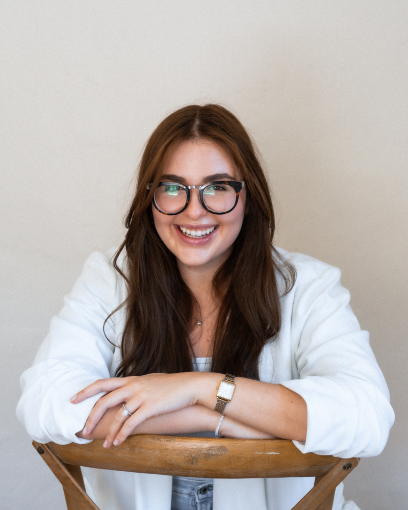 Portrait of Rafa, the lead designer of Alella Creative Studio, smiling warmly at the camera. She wears glasses and a white blazer, with her hands clasped on a wooden surface.