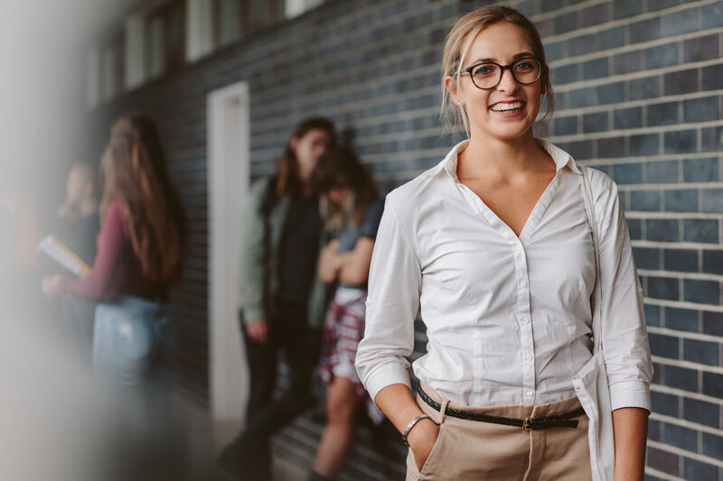 A white woman assistant professor walks outside an academic building. She is smiling with one hand in her pocket and a canvas bag on her left shoulder. In the background, two students lean against a brick wall chatting.