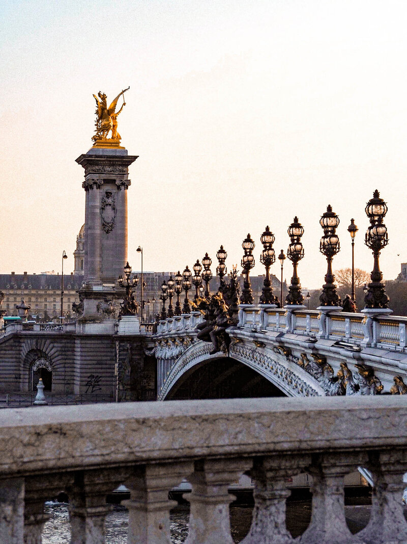 Ornate bridge with intricate lamp posts and golden statues at sunset, echoed in the elegance of https://epicsandstravel.com/about. The bridge spans a river, with a gold-topped column featuring a winged figure in the background against a light pink and orange sky.