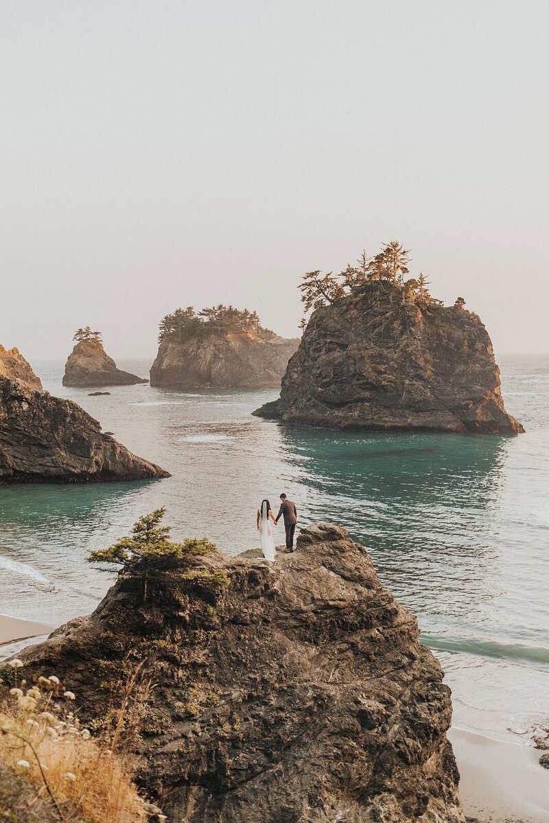 A light ray shines through an arch rock onto a couple in wedding attire on the Oregon Coast