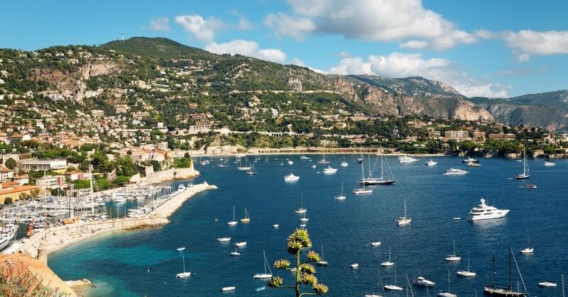 View of the Bay of Nice, featuring sailboats gliding over azure waters and the scenic Promenade des Anglais in the French Riviera.