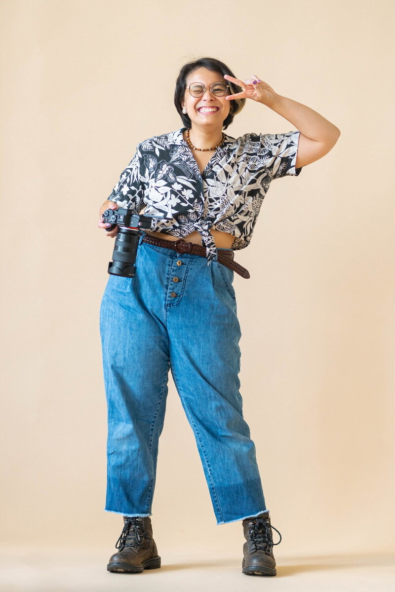 Erin Deleon, holding her camera and throwing the peace sign in front of a beige background