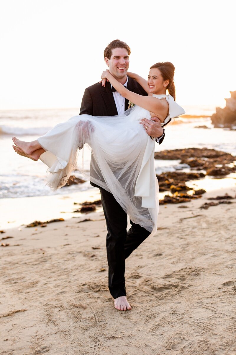 groom carries stylish bride along orange county beach