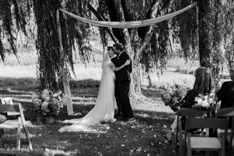 A bride and groom share a kiss under a simple arch adorned with fabric, standing in front of a large tree. Captured by a documentary photographer in New England, they are outdoors, surrounded by guests seated on chairs with floral arrangements nearby, conveying a serene, intimate wedding setting.