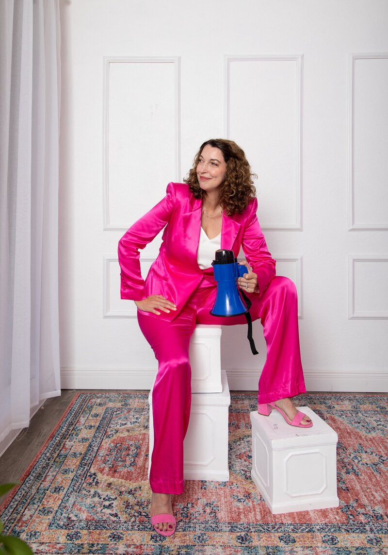 Amanda Hennessey, public speaking coach, in bright pink suit holding megaphone, sitting confidently, smiling
