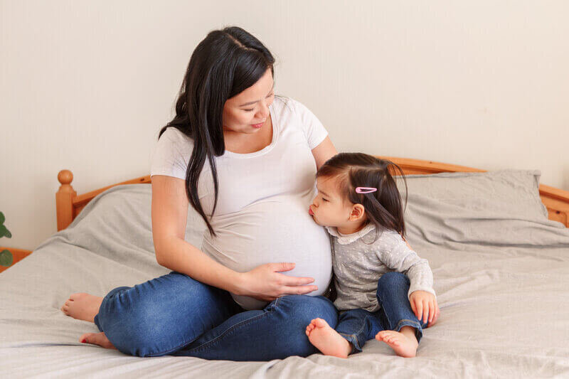 Pregnant woman and child sitting on bed, child is kissing the woman's pregnant stomach