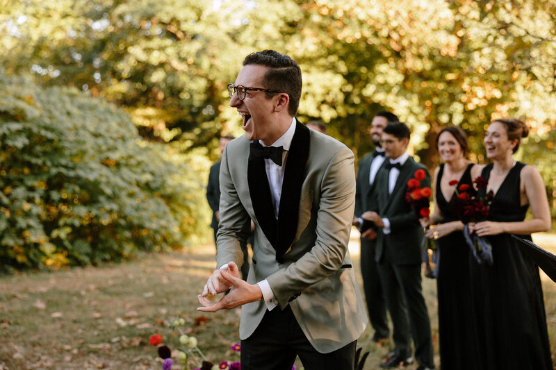 An exuberant groom gestures expressively while standing among elegantly dressed guests at an outdoor gathering, surrounded by lush greenery.