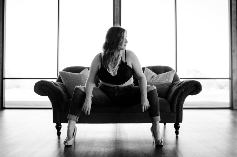 black and white boudoir photo with woman in a blue jeans and a black bra sits on a couch and looks off to the side as she rests her elbows on her knees at little rock ar boudoir studio