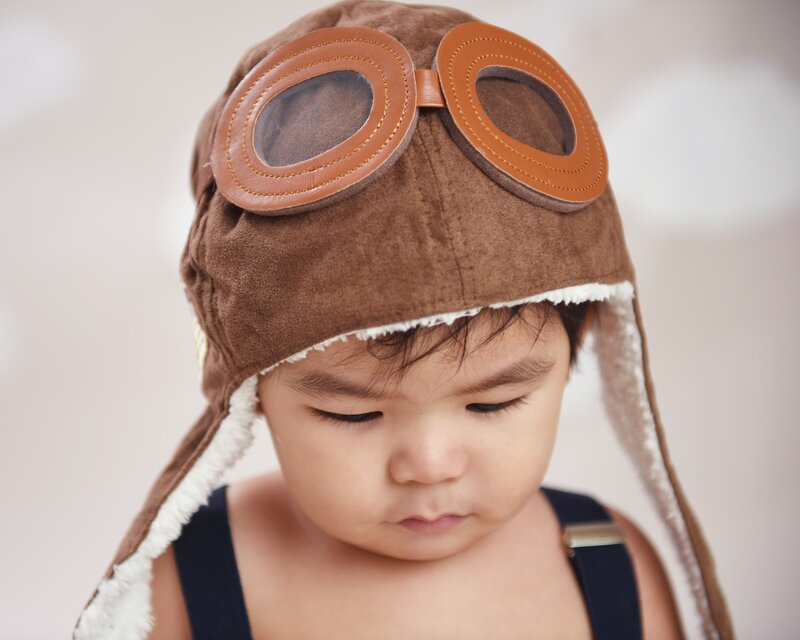 A close-up of a baby boy wearing a brown aviator hat with goggles, looking down, in a soft and dreamy backdrop.