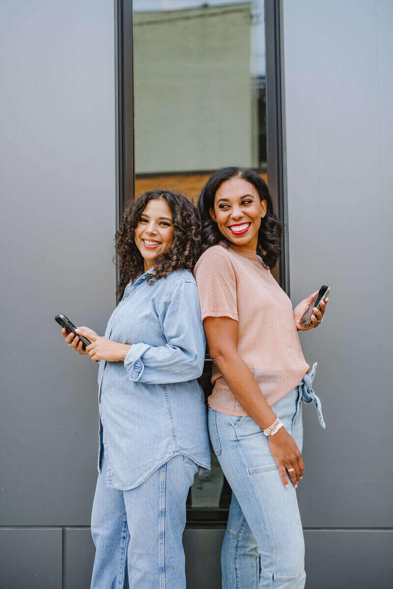 Two woman smiling standing back to back with phones in hand.