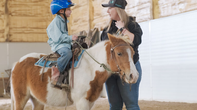 Spirit's Respite Ranch's Coach guiding a child in horseback riding, creating a nurturing connection between the child and the horse in the serene Alberta countryside.