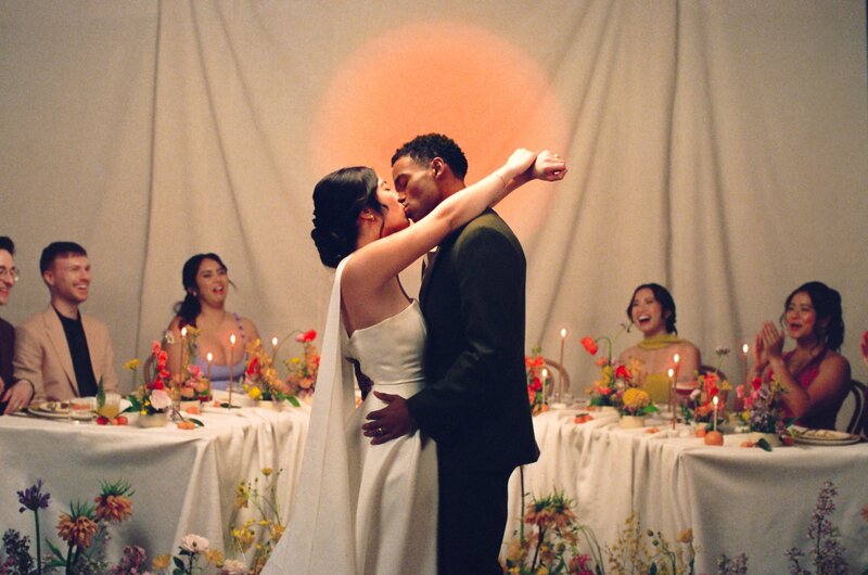 Film photo of a bride and groom kissing in front of a round sunset backdrop during a candlelit wedding dinner, surrounded by joyful guests — a fine art wedding moment by a film wedding photographer.