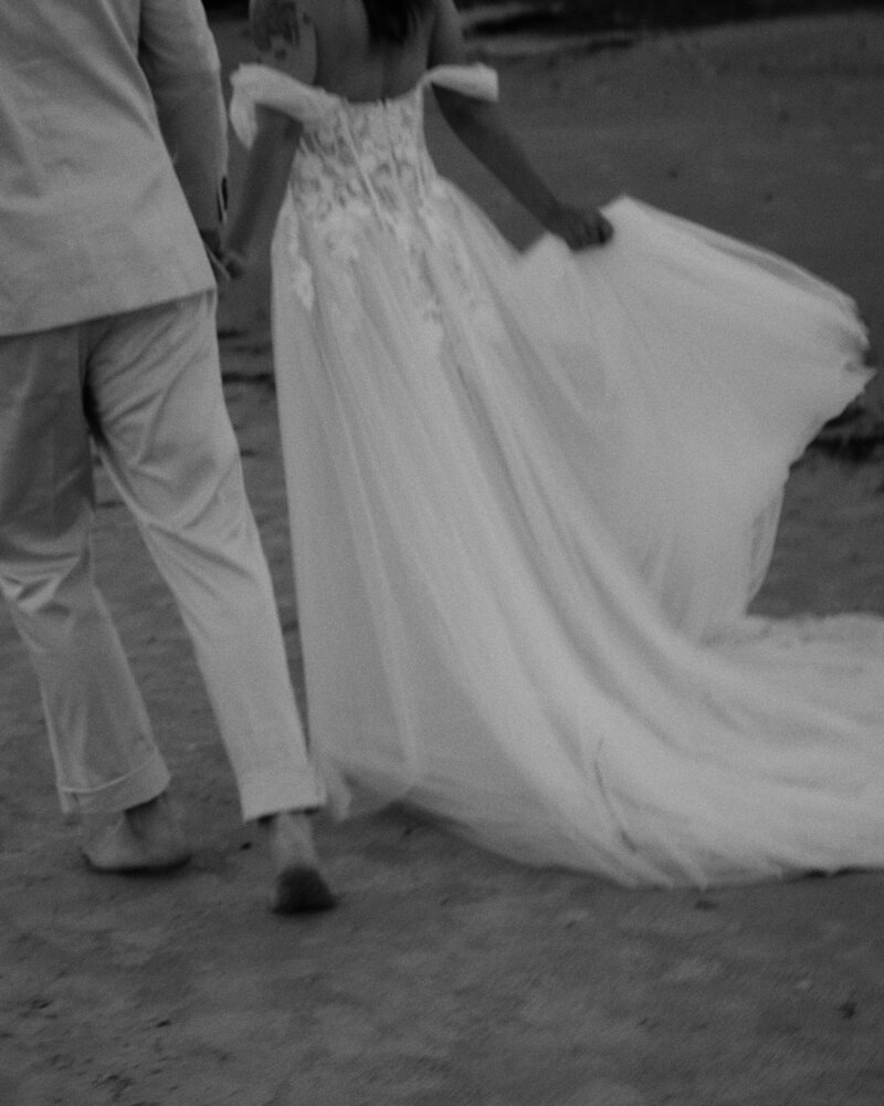 Wedding dress flowing while couple walks on the beach