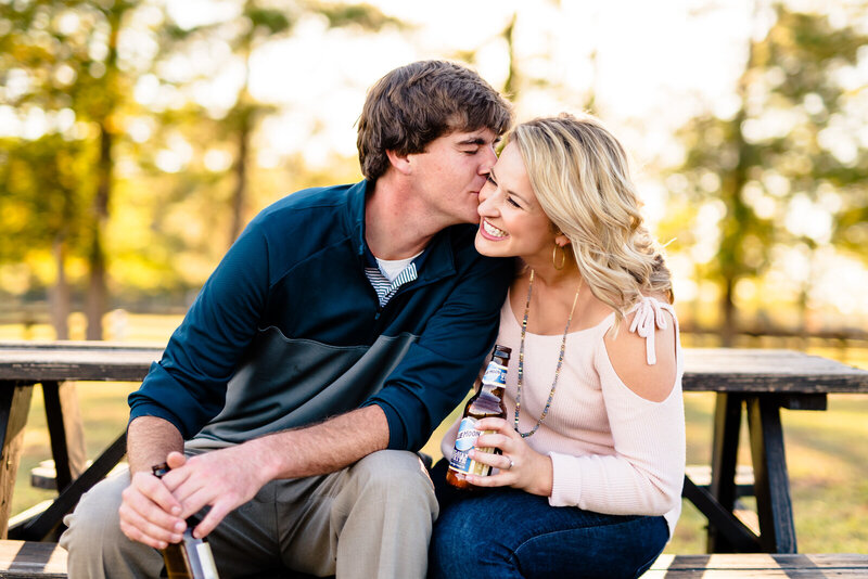 Man kisses woman on the cheek while sitting at a picnic table during an engagement photo session