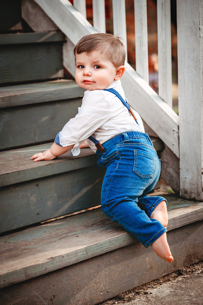 A one-year-old boy in suspenders is climbing up wooden stairs at his baby photoshoot.