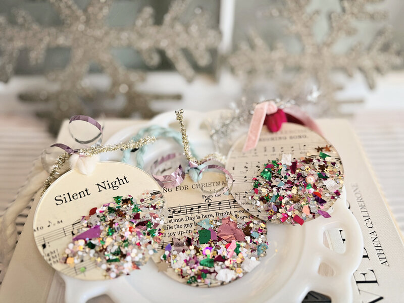 Handmade song-themed acrylic ornaments by Sweet Pea Ink Creative, featuring vintage sheet music designs, festive glitter confetti, and decorative yarn and ribbon. Shown on a white plate with glittery snowflakes in the background.
