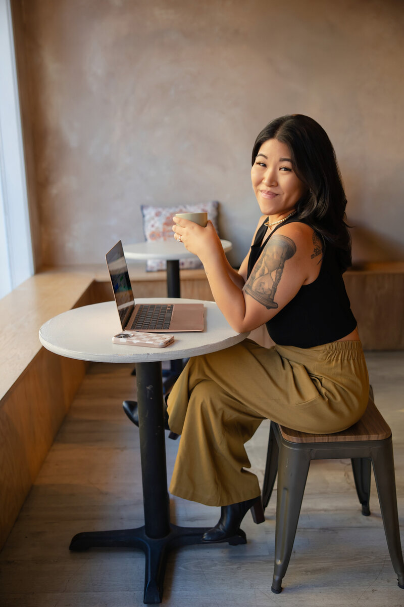 Woman with long black hair and visible tattoos sits at a café table with a laptop and coffee cup, smiling confidently over her shoulder. Captured during a personal branding photoshoot, this image highlights a relaxed yet empowered vibe perfect for creative entrepreneurs and coaches.