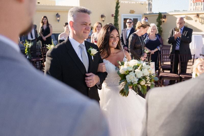 A bride in a white strapless wedding dress walks down the aisle arm-in-arm with her father in a black tuxedo during an outdoor wedding ceremony at the White Room in historic downtown St. Augustine. She carries a white and green bouquet while wedding guests look on from their seats in the background. The venue features elegant architecture with arched doorways and outdoor lighting.