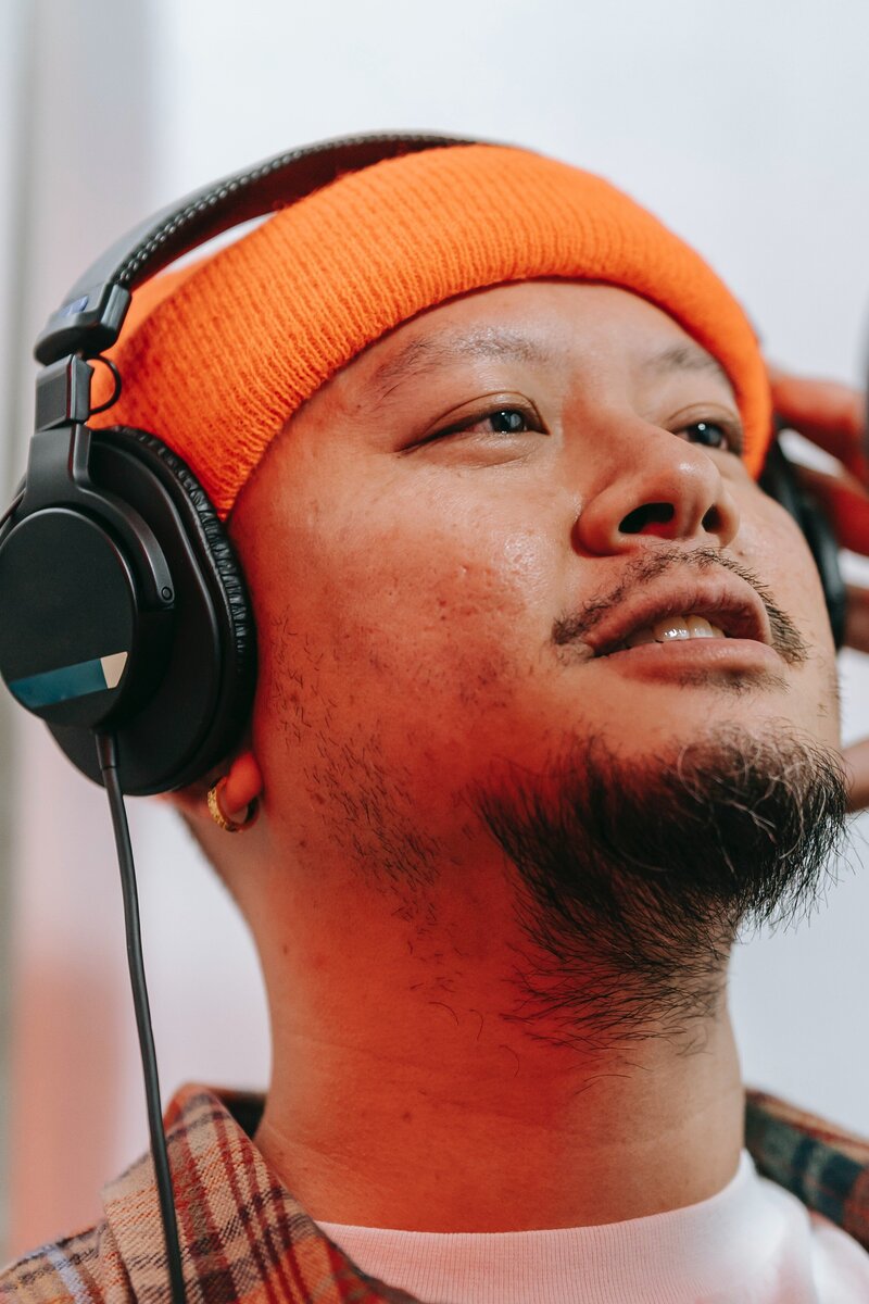 This image shows a close-up view of a masculine-presenting Asian individual, wearing a beanie and headphone, listening to music. They are looking up toward the sky, past the camera.