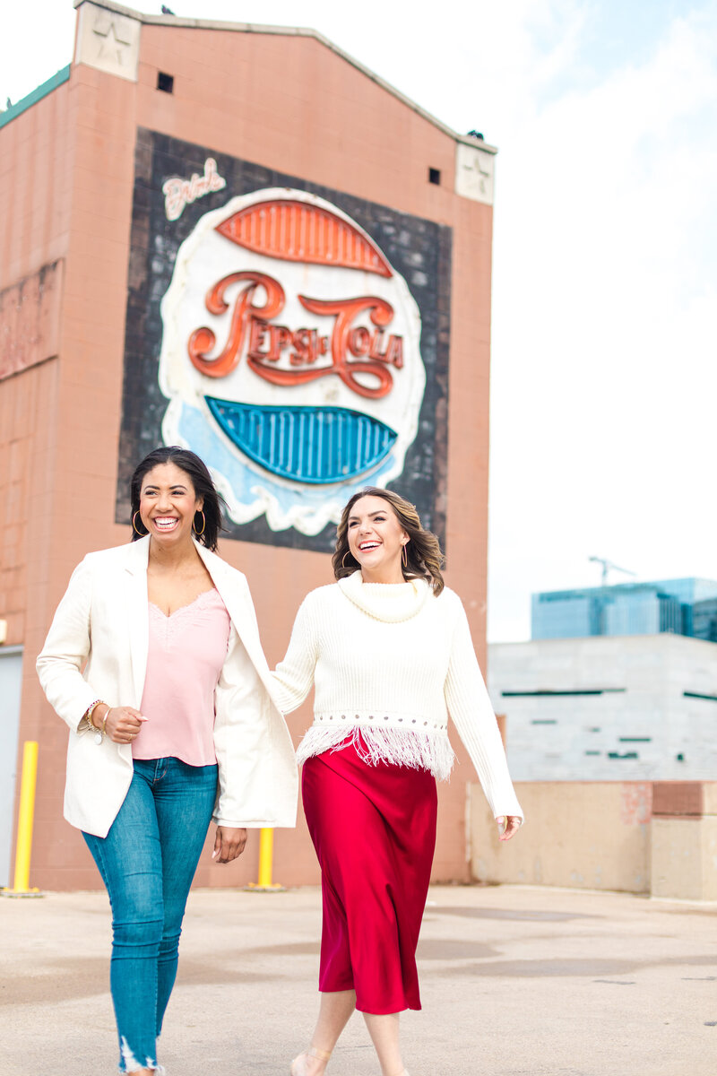 Two women are walking in front of a terracotta building with a giant black, white and red sign as an advertisement. The women are smiling and one of them is wearing red pants with a white blazer and the other woman is wearing jeans with a light pink blouse.