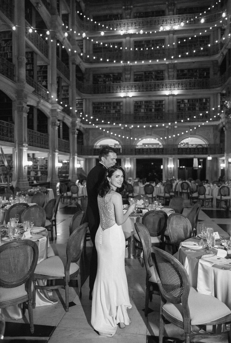 A black-and-white photo of a bride and groom standing in an elegantly decorated dining hall. The room features high shelves filled with books and string lights hanging from the ceiling. The bride wears a long gown, holding the grooms arm.