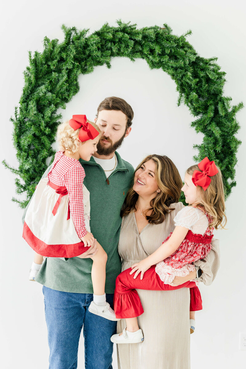 Family of 4 posing together smiling at each other in front of wreath for christmas mini sessions