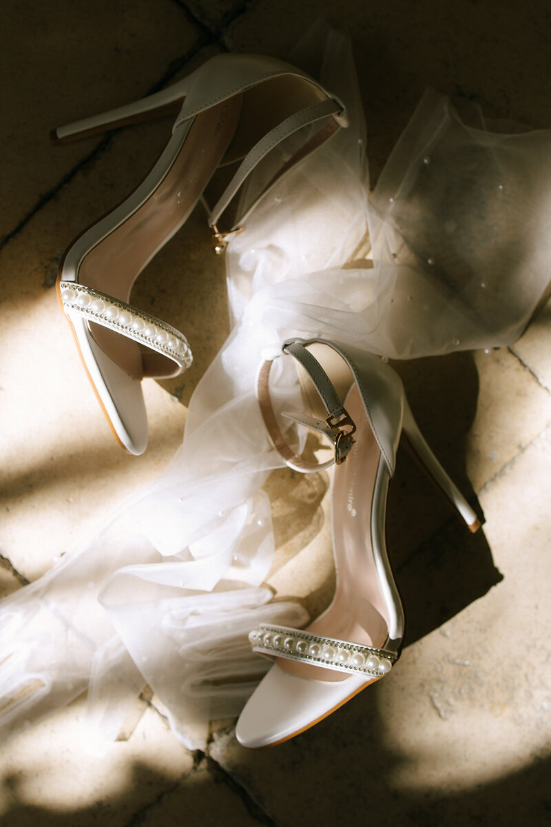 A pair of elegant white bridal heels adorned with pearls, styled on soft tulle fabric and bathed in natural light