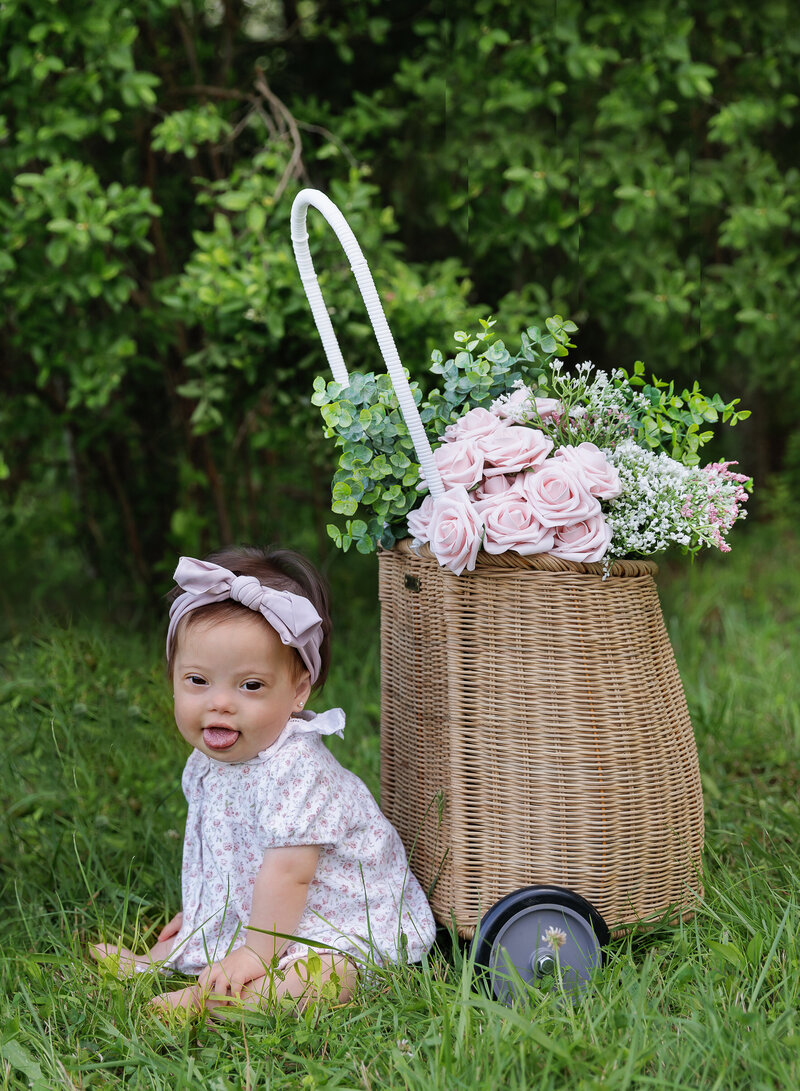 Baby girl sits on the grass  for her first birthday photoshoot. She is looking over her shoulder and smiling at the camera.  Captured by best Brooklyn, NY milestone photographer Chaya Bornstein.