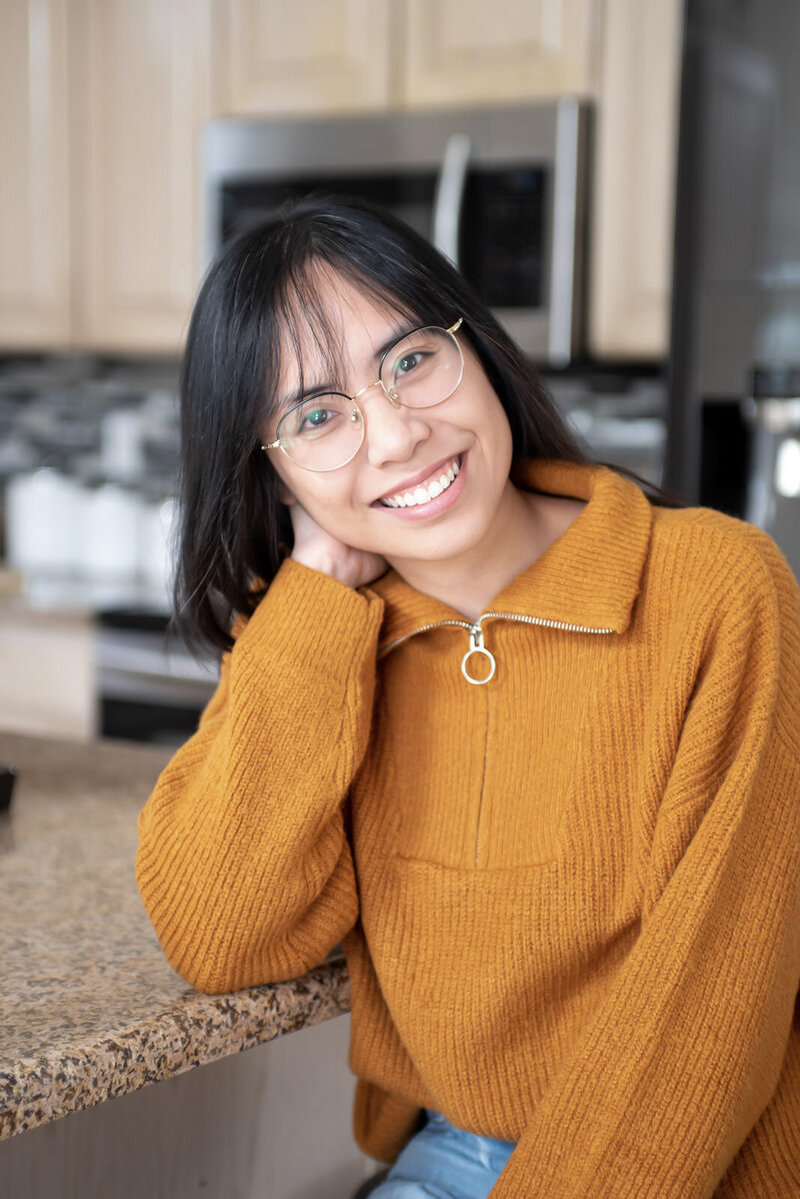 Laci smiles while resting her head in her hand and wearing a mustard colored sweater