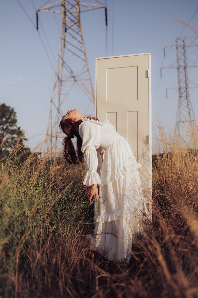 Woman in ruffled white gown stands in grassy field with powerlines and poses standing with her back arched in front of a white door.