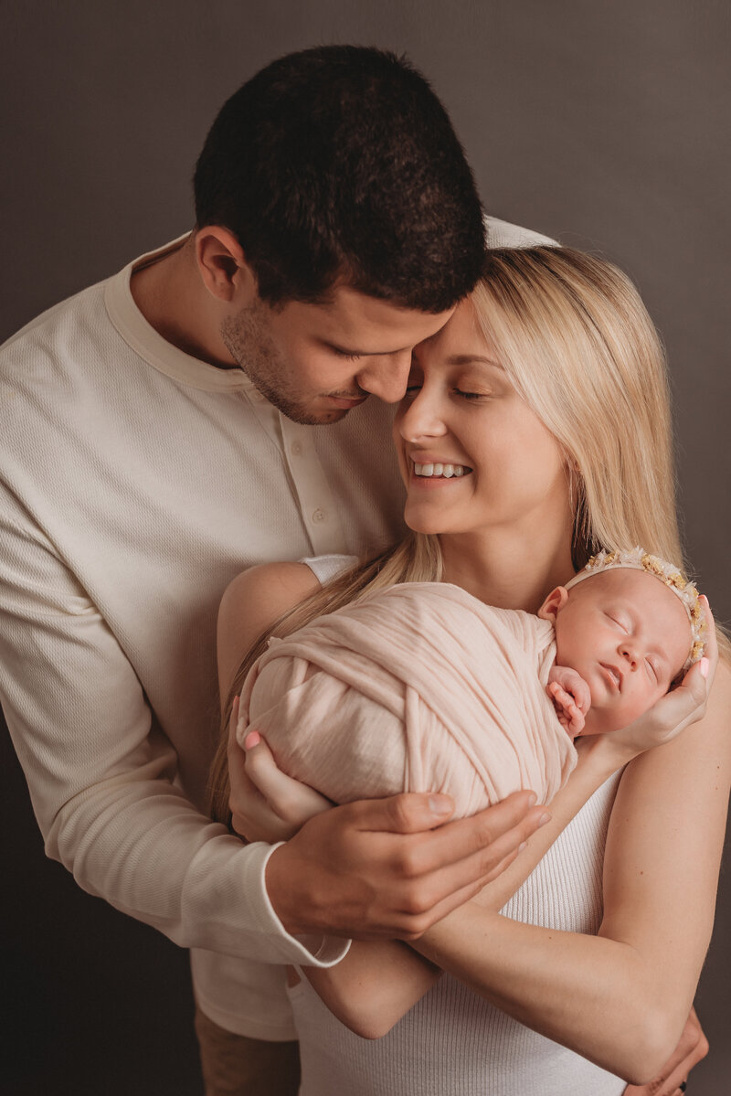 Family of three standing up with mom holding newborn baby girl wrapped in light pink swaddle and mom and dad are nose to nose smiling