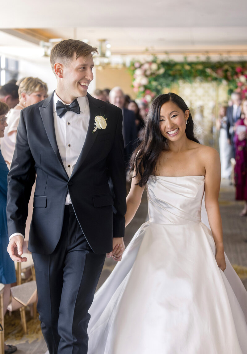 A smiling bride and groom walk down the aisle. The bride wears a strapless white gown, and the groom wears a black tuxedo with a bow tie. They hold hands, with wedding guests and floral decorations visible in the background.