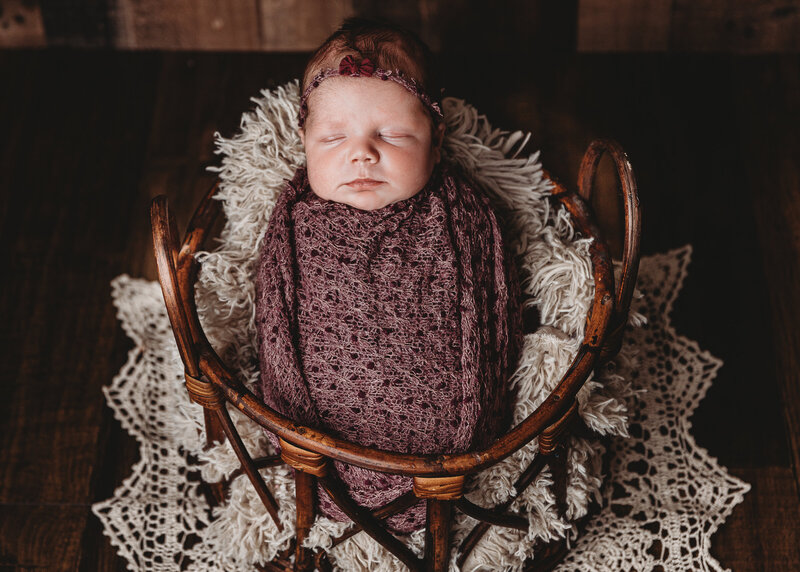 A baby swaddled in a lavender blanket, place in a basket.