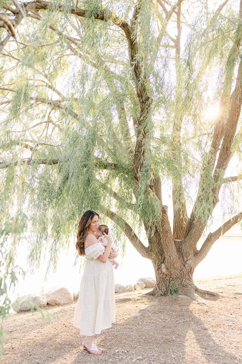 mother standing in front of a willow tree at sunset holds her newborn close to her chest in a cream dress,  Indianapolis newborn photographer