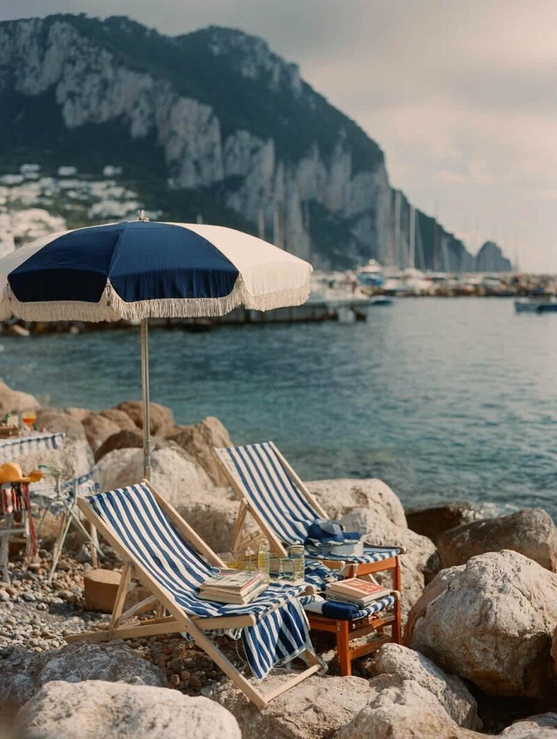 Seaside retreat with striped lounge chairs and umbrella perched above turquoise coastal waters in Europe. Tailored Oasis Travel Co.
