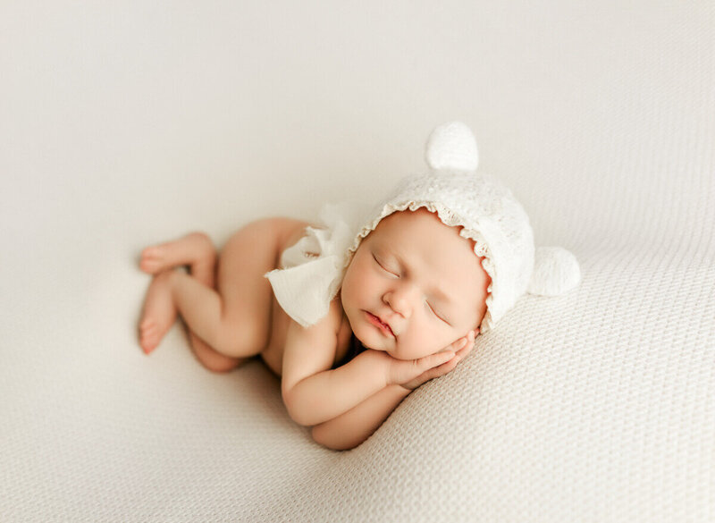Newborn baby sleeping on their side with hands tucked under chin, styled with neutral fabrics in a fine-art studio setting.