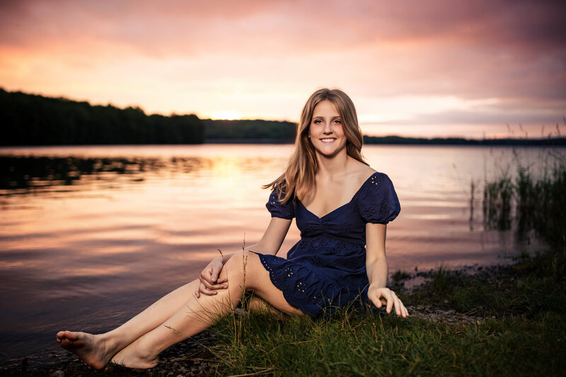  This blond senior girl poses against a bright pink sky and a reflective lake.