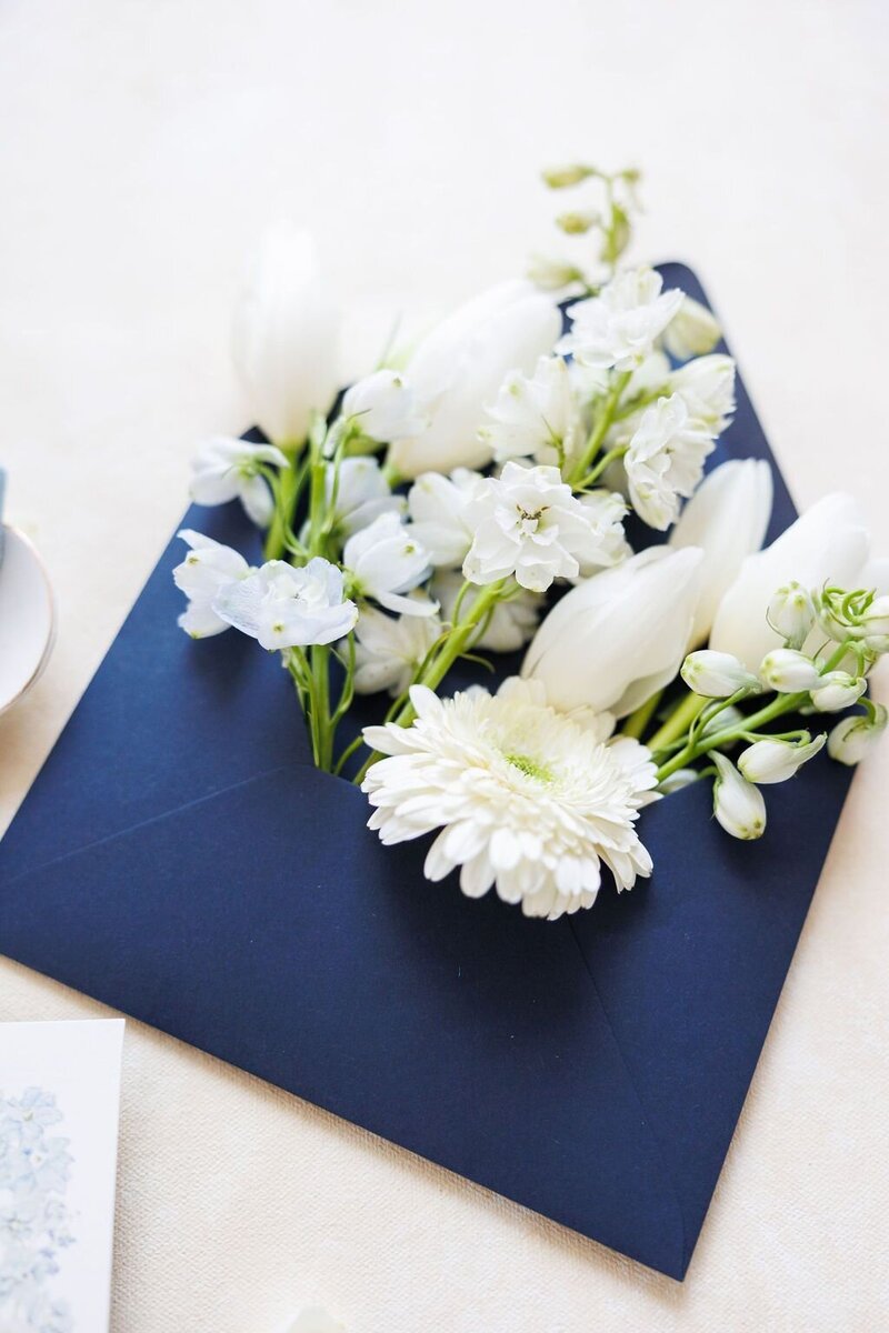 White florals neatly tucked into a navy paper envelope.  Flat lay, invitation