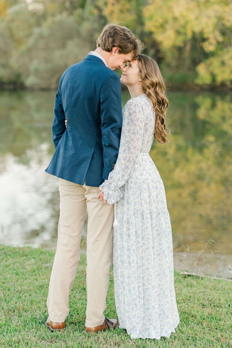 Engaged couple with backs to camera holding hands and looking into each others eyes inf ront of water