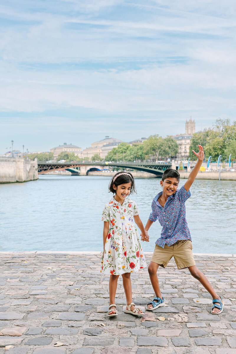Young boy and girl holding hands, with the boy raising his arm in a playful pose during a Paris family photoshoot on Île Saint-Louis in the city center.