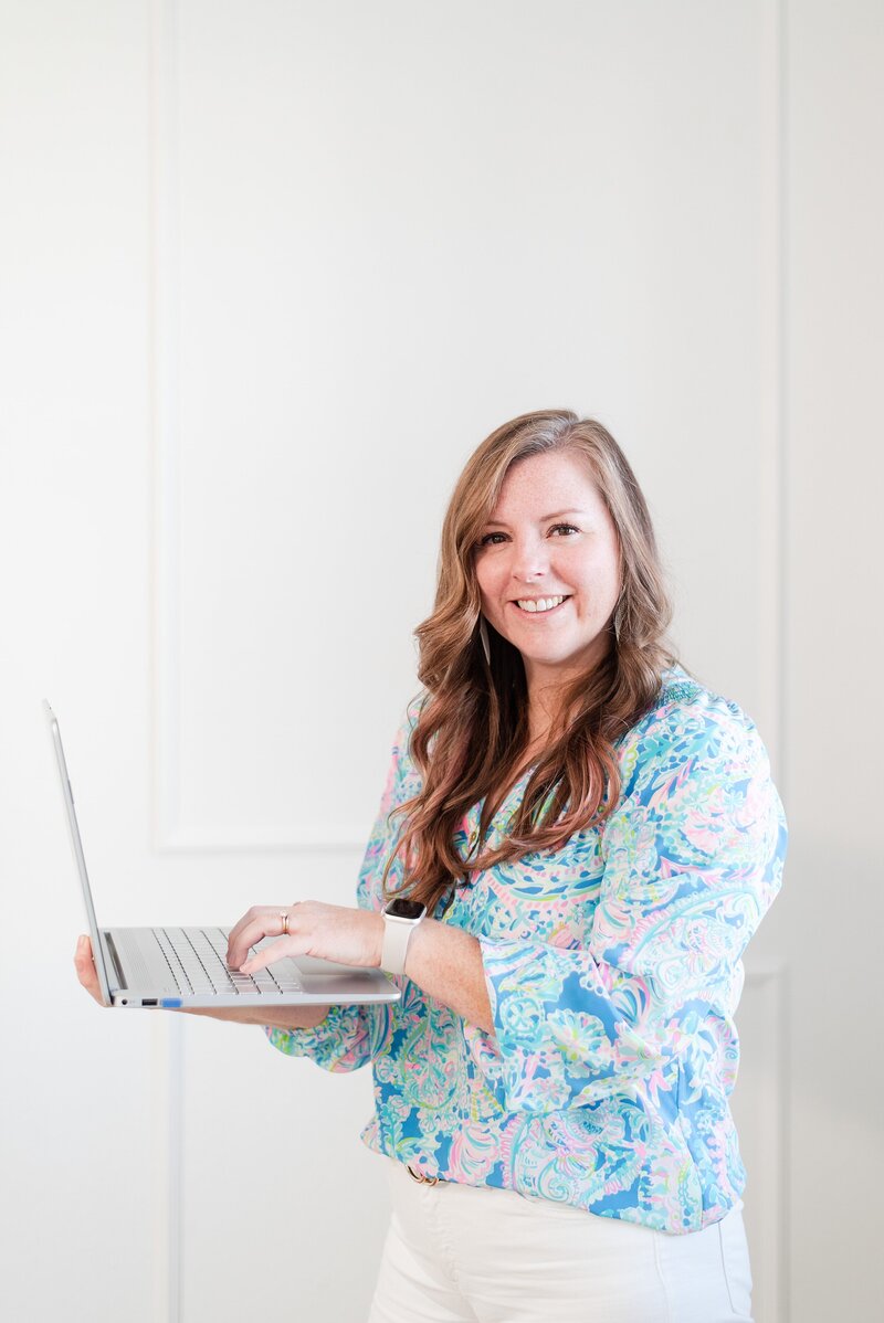 melissa a photographer seo mentor smiling at camera holding her laptop  while wearing a brightly colored lilly pulitzer shirt and her long brown hair is curled
