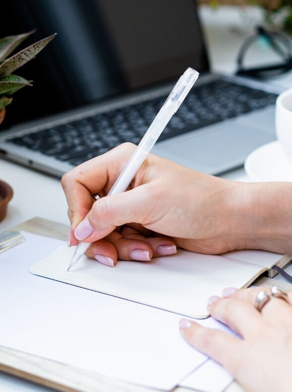 Woman's hand with pink nails writing something in a planner with laptop in background