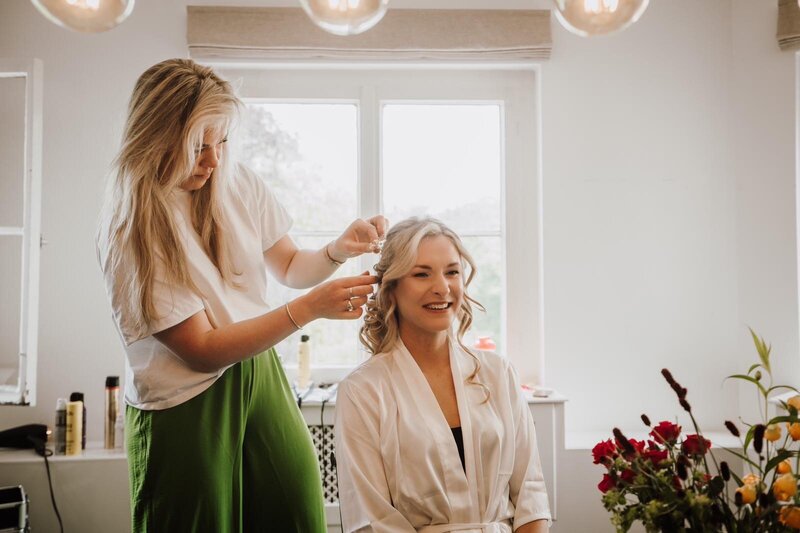 Een bruidsstylist bevestigt een haaraccessoire in het opgestoken kapsel van een glimlachende bruid. De bruid draagt een witte satijnen ochtendjas en zit in een lichte kamer met een groot raam. Op de voorgrond staan rode bloemen.