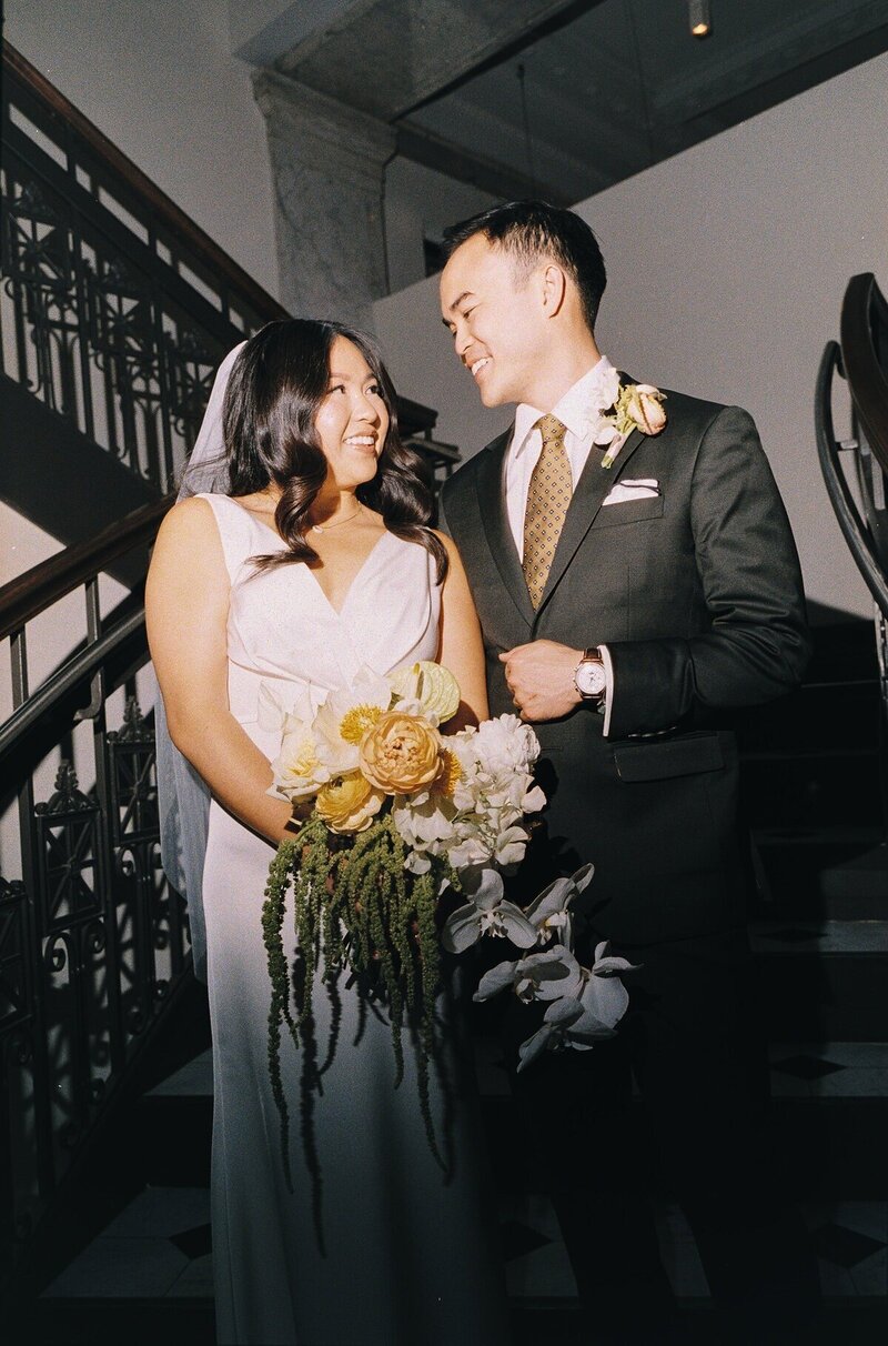 image by tom thornton photography of bride and groom on wedding day holding bouquet standing on staircase smiling at each other 