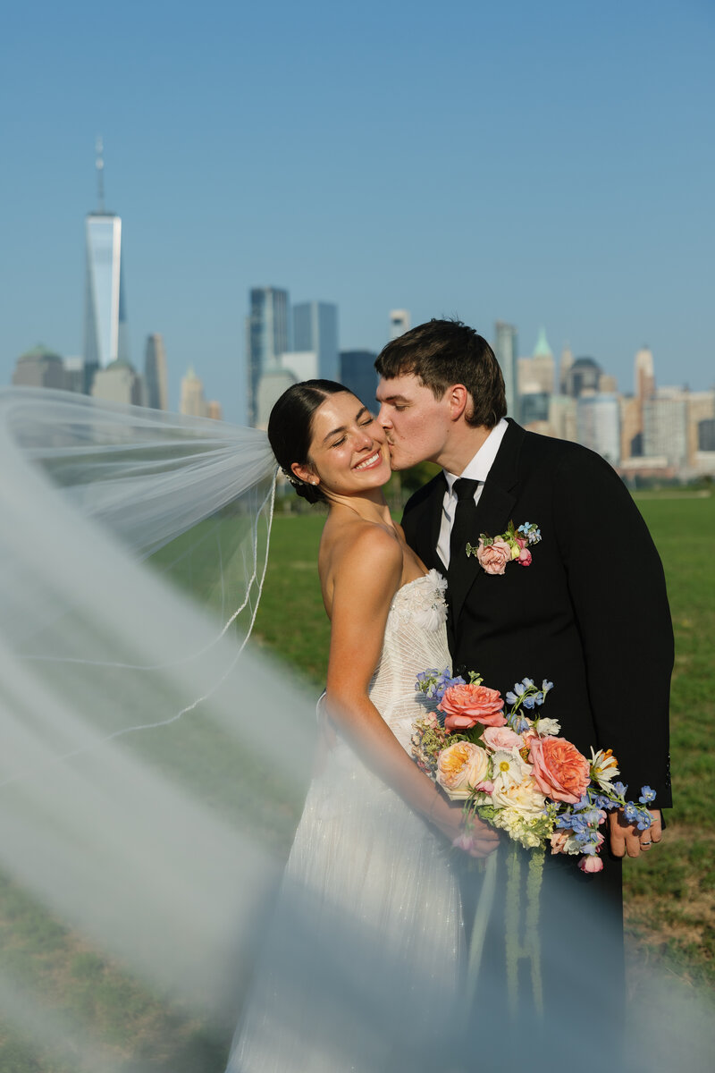 A bride and groom share a kiss on a sunny day, with a flowing veil and a bouquet of colorful flowers against NYC skyline backdrop.
