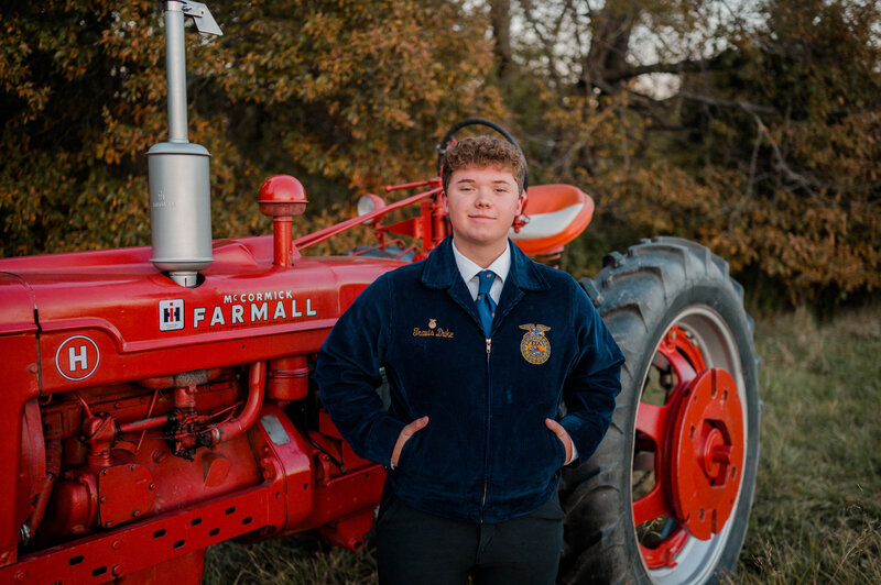 A high school senior boy poses for the camera in his FFA jacket and leans against a red tractor.