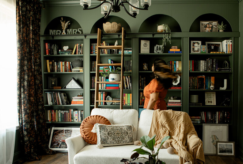 A woman in an orange dress walks through a green library with a rolling ladder. There's a white couch in front of her and plants all around.