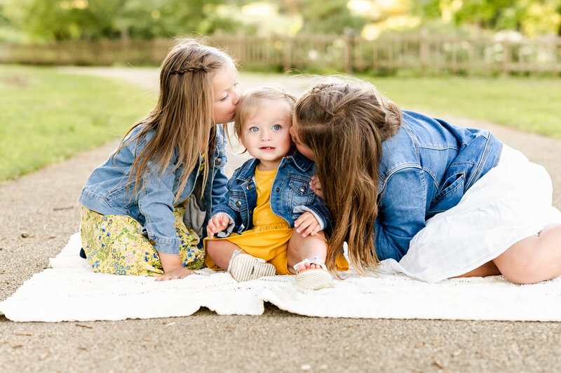 Older sisters kissing youngest while on a blanket at Chicago family photography session.