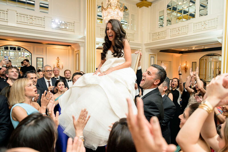 A bride in a white dress is joyfully lifted on a chair during a celebration. She is surrounded by smiling guests in formal attire, including a man holding the chair. The room is elegantly decorated with columns and a chandelier.