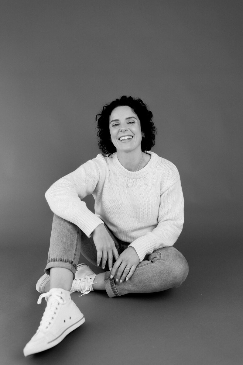 Nicole gourley, christchurch wedding photographer, sit on floor in white sweater and jeans in posed studio portrait